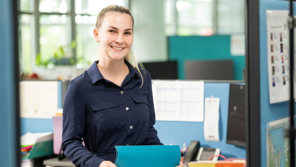 Woman stands in an office holding a blue folder and smiling warmly.
